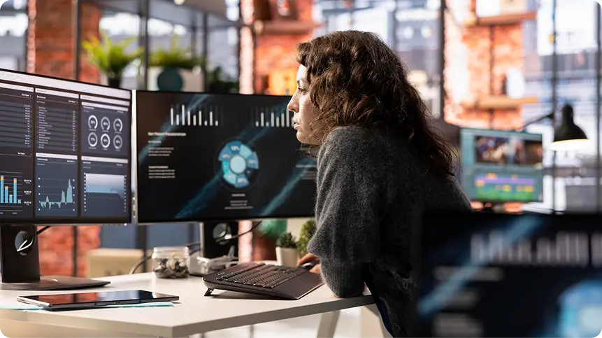 Woman at a three-monitor workstation showing dark dashboards with charts and data panels