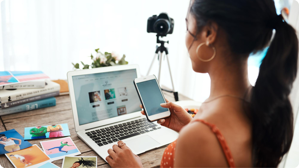 Creadora de contenido revisa su teléfono frente a una laptop con un blog y una cámara sobre trípode